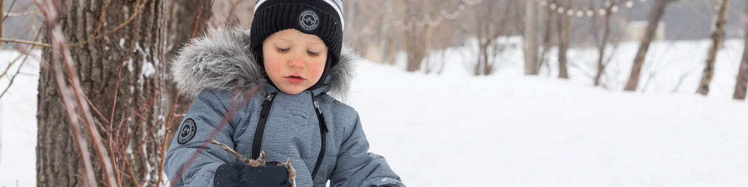 un bébé garçon habillé avec la marque Pluche et tartine assit dans la neige sur le bord d'un arbre