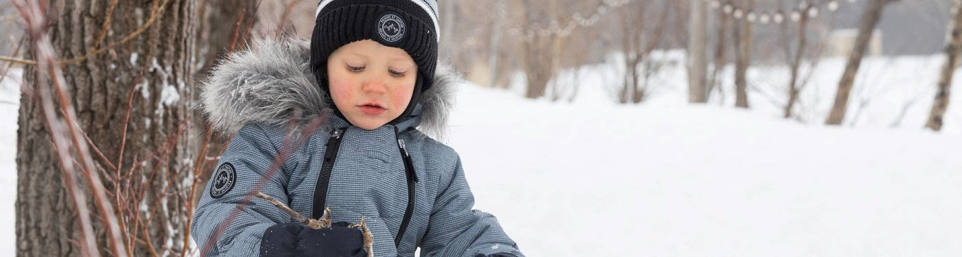 un bébé garçon habillé avec la marque Pluche et tartine assit dans la neige sur le bord d'un arbre