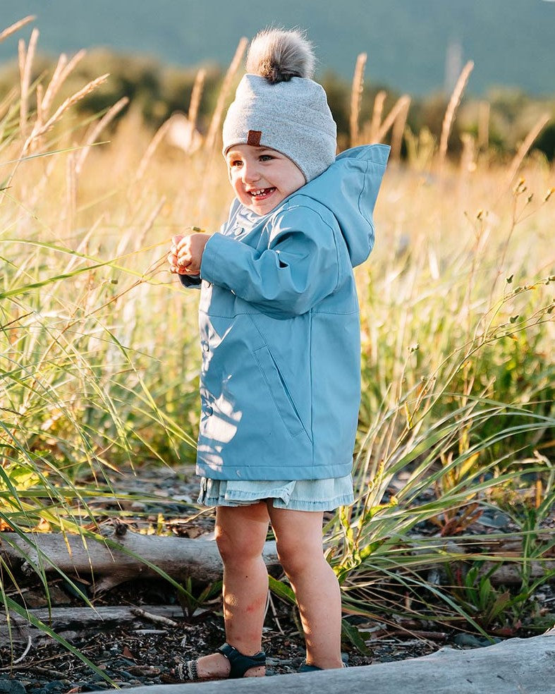 une jeune enfant avec une tuque grise à pompom et un coupe-vent mi-saison bleu sur le bord de l'eau